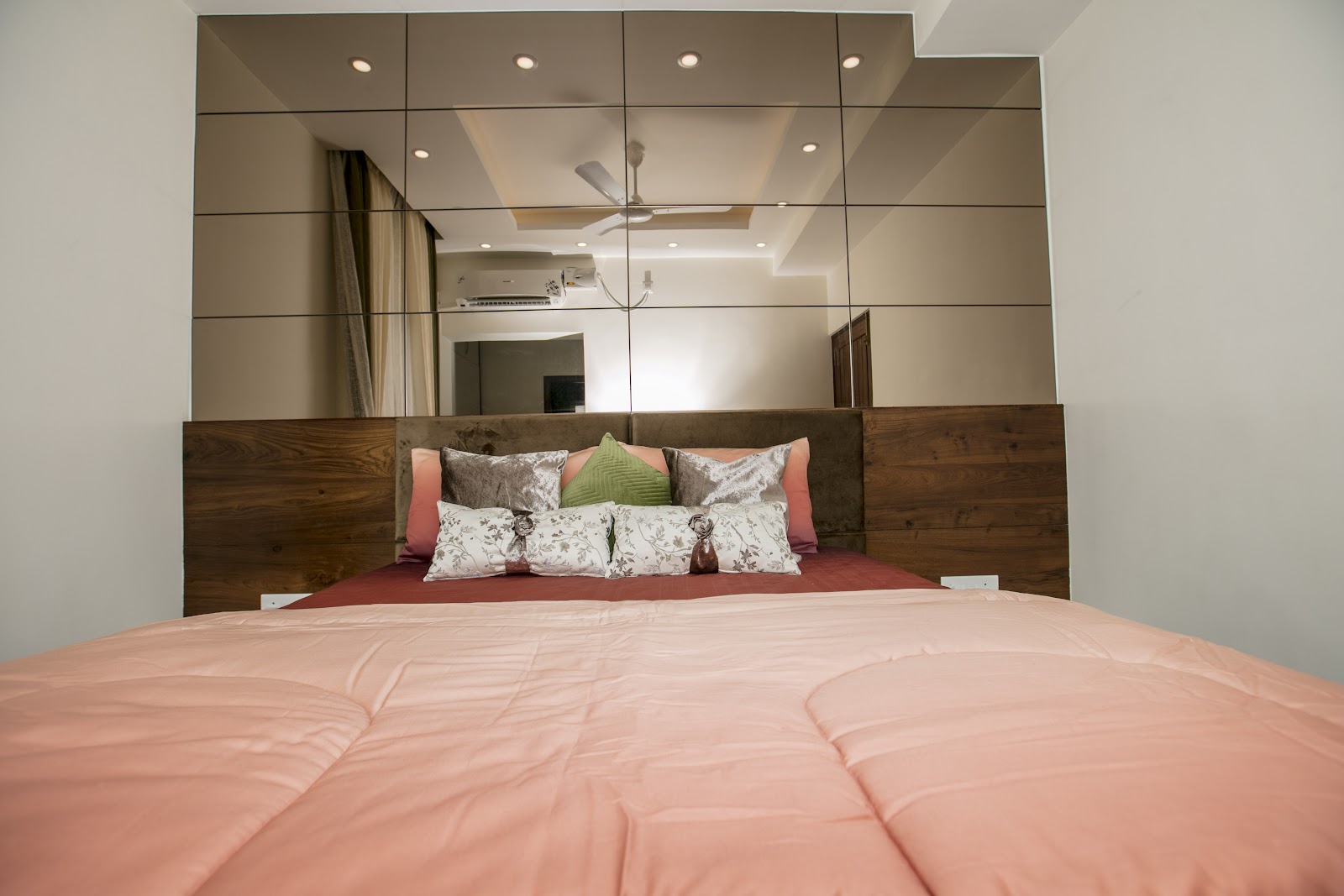 A wood-and-upholstered headboard bed with coral bedding, set beneath a full mirrored panel wall that reflects the ceiling and fan.
