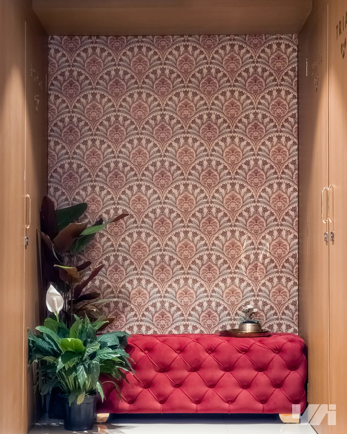 Entrance foyer nook with a burgundy damask wallpaper wall, a red tufted velvet bench, and potted plants, framed by open wardrobe doors on either side.