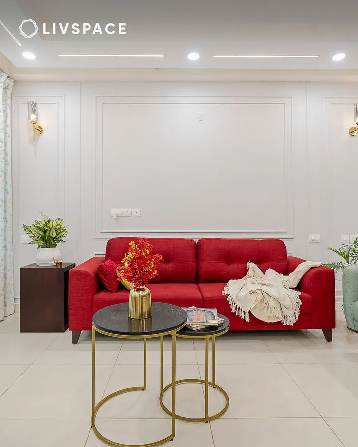 Red fabric three-seater set against a white moulded panel wall flanked by brass cylinder sconces, with nesting black-marble-top brass tables on ivory tiles.