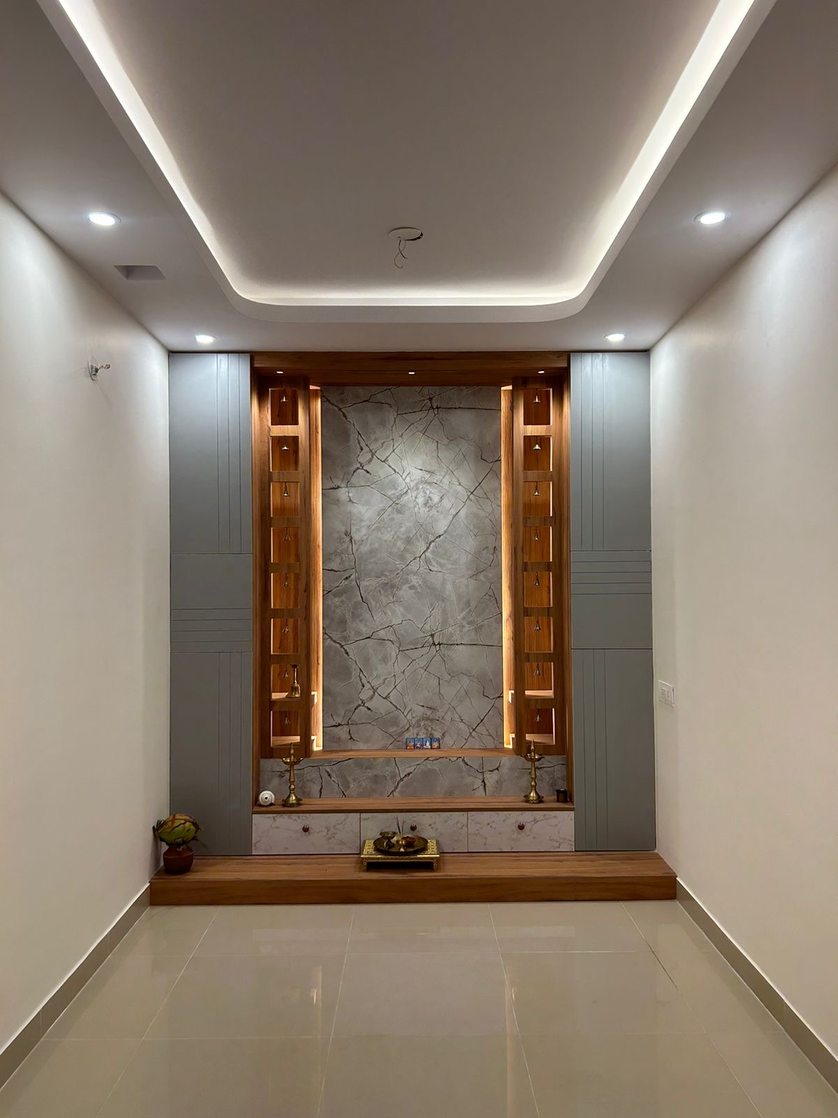 Dedicated pooja alcove with a crackled grey marble backdrop framed by wood display niches holding brass bells and diyas, under a cove-lit ceiling.