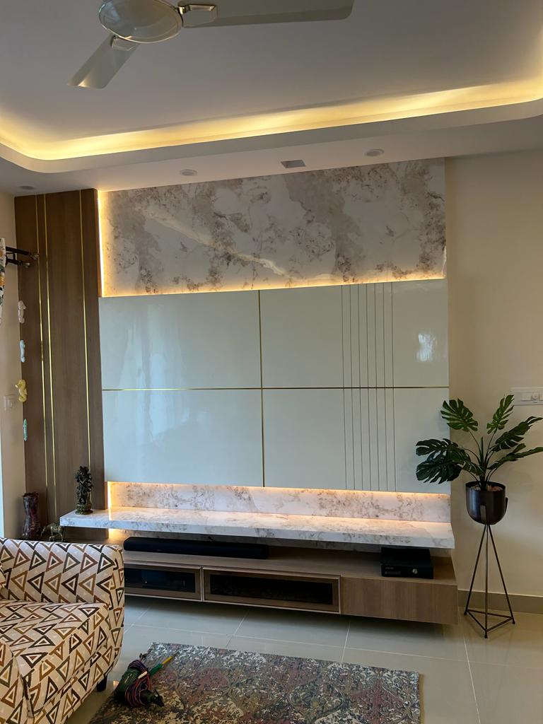 Living room TV wall with marble-clad top and bottom bands framing a glossy gold-lined panel, beside a fluted wood pillar and a monstera plant on a metal stand.