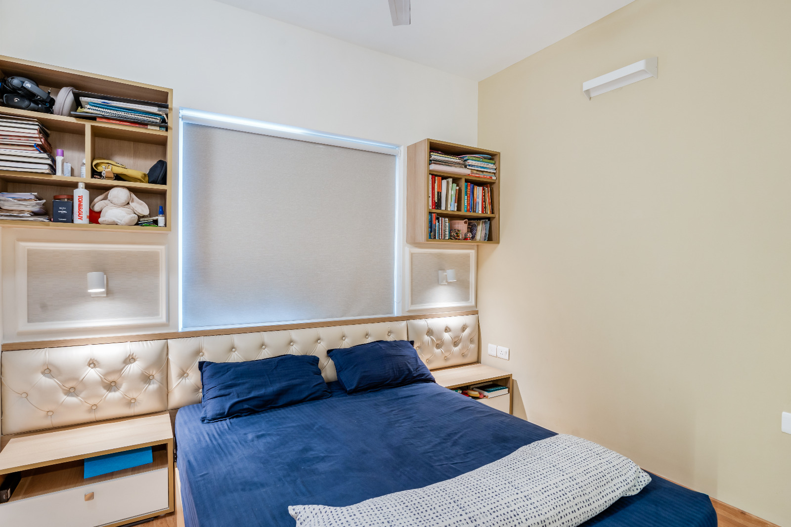 A bedroom with a cream diamond-tufted upholstered headboard flanked by wall-mounted bookshelves and sconce reading lights, and a navy bed cover.