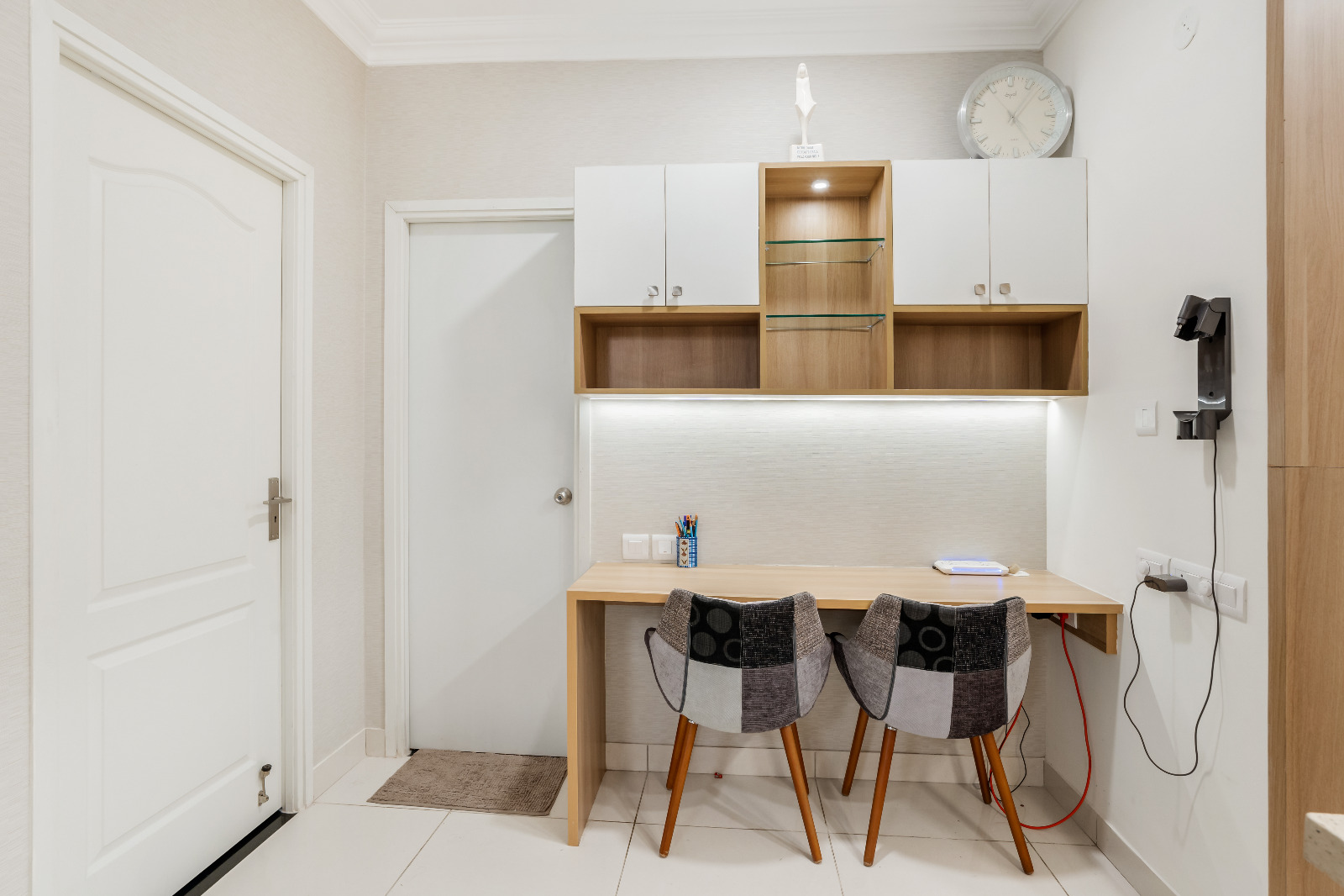 Home study nook with a built-in wood and white desk unit featuring a glass-shelf display niche, LED-lit backsplash, and two patchwork upholstered chairs.