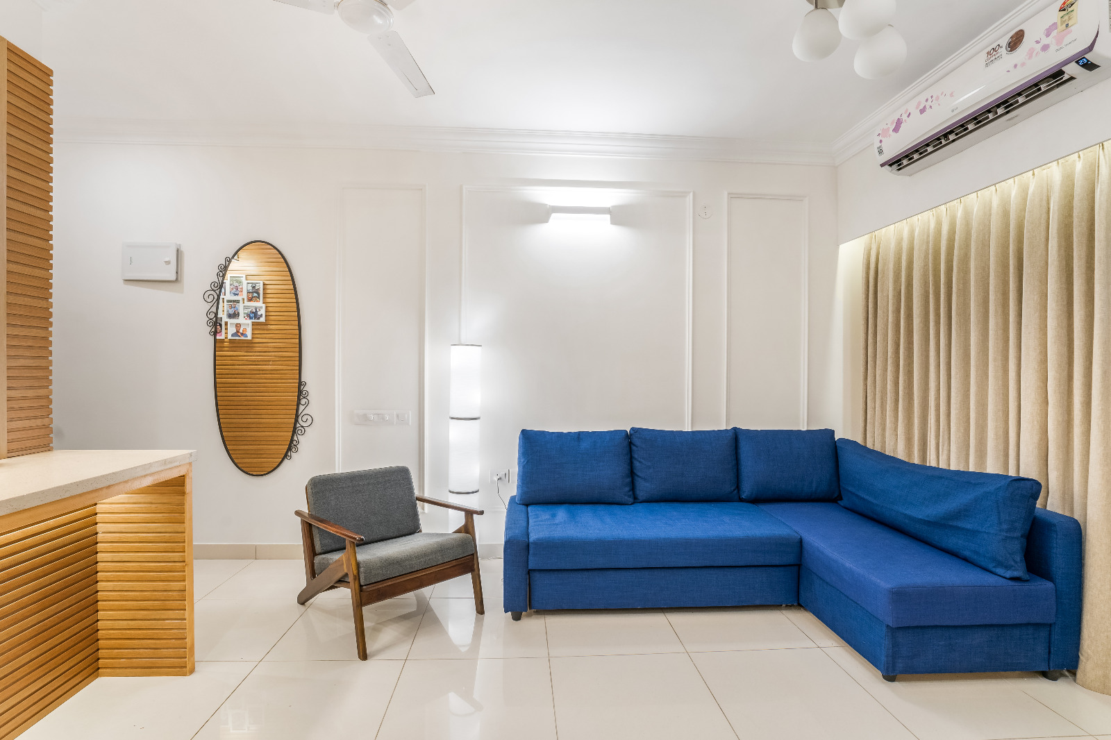 Living room with a royal blue L-shaped sofa, a mid-century wood-frame armchair and an oval mirror with a family photo collage.