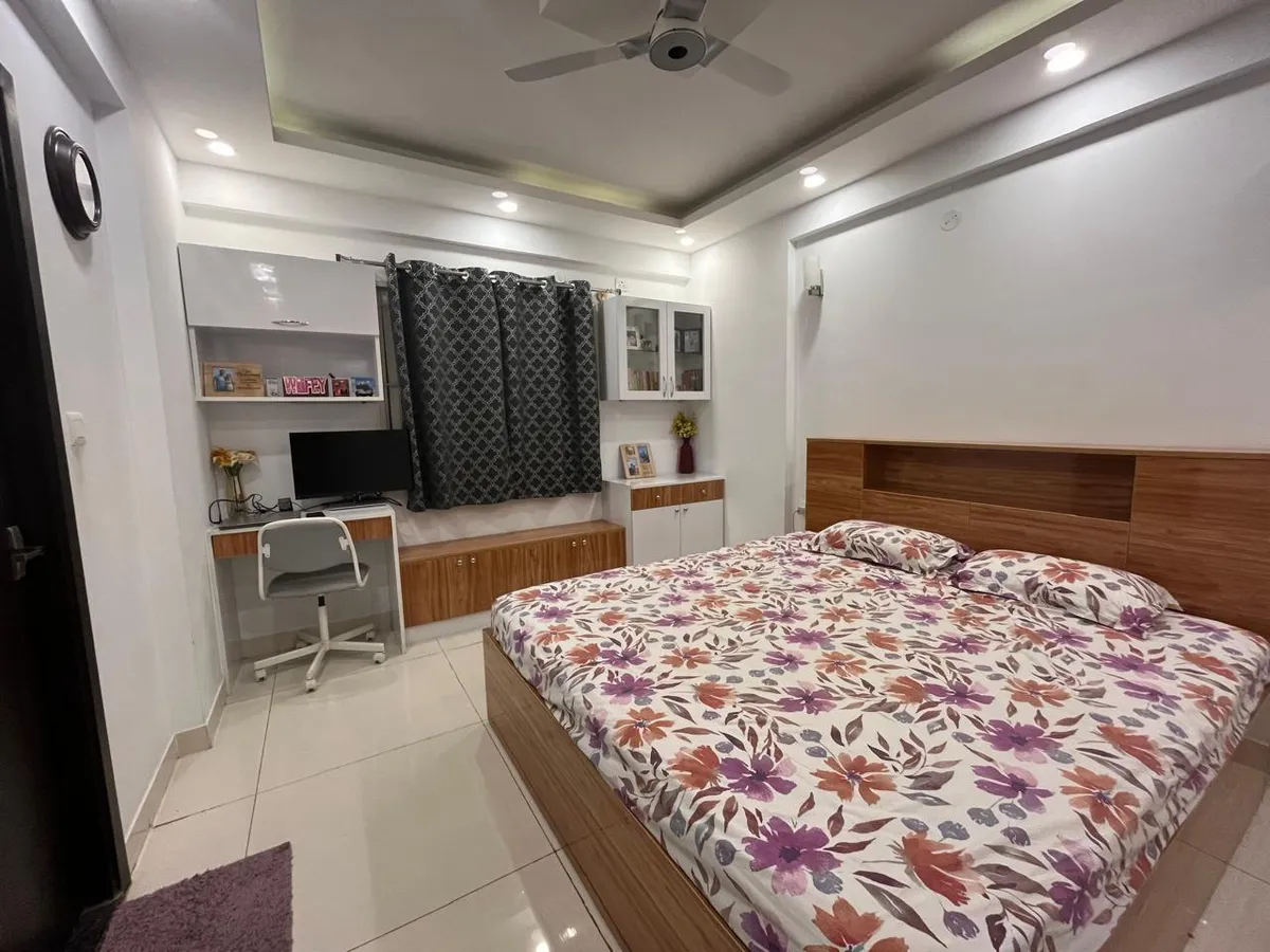 Compact bedroom where a teak-laminate headboard with a built-in display ledge meets a white-and-teak study desk under a gloss wall cabinet