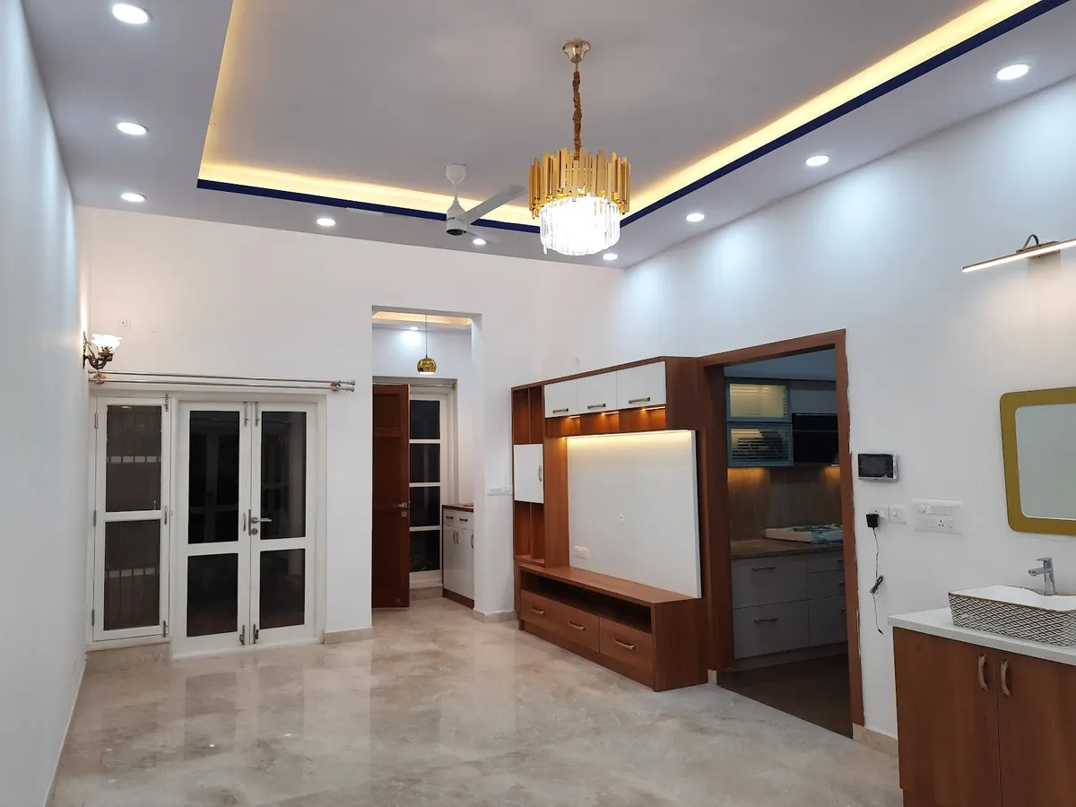 Real photo of an open-plan living space with a wood-and-white TV unit, a crystal-and-gold chandelier under a cove-lit ceiling, and glass French doors
