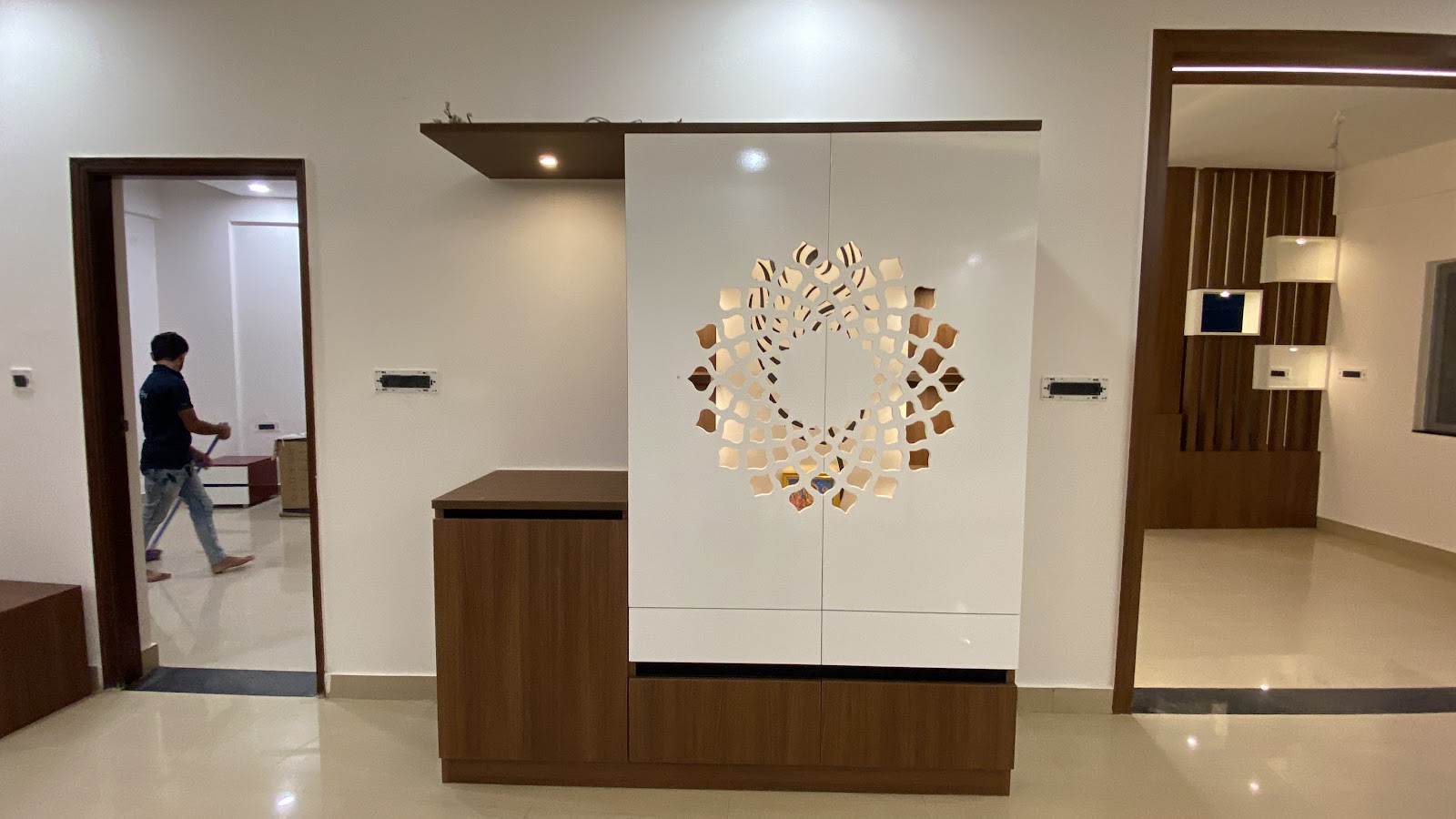 A white lacquered pooja cabinet with a backlit laser-cut mandala medallion, paired with a walnut console, in an entryway opening to other rooms.