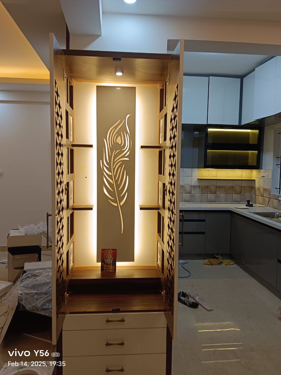 Free-standing pooja unit with jaali-patterned side doors and a backlit wood panel carved with a peacock-feather motif, drawer storage below