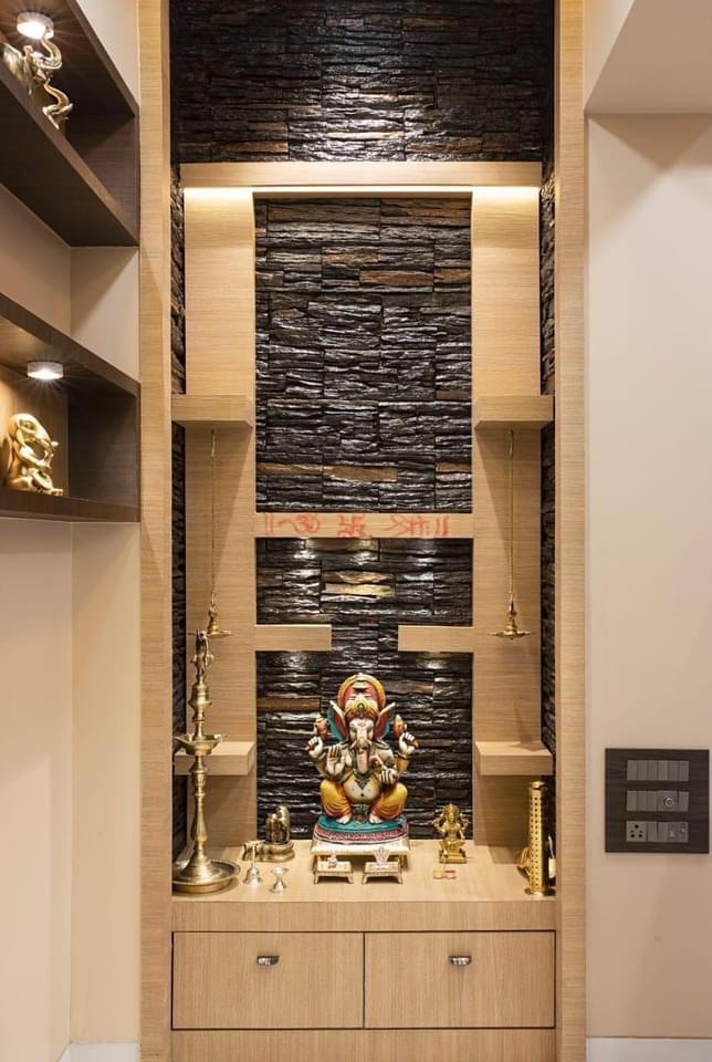 Pooja niche with dark stacked-stone cladding, wood shelving flanked by hanging brass diyas, a Ganesh idol on the counter, and drawer storage below.