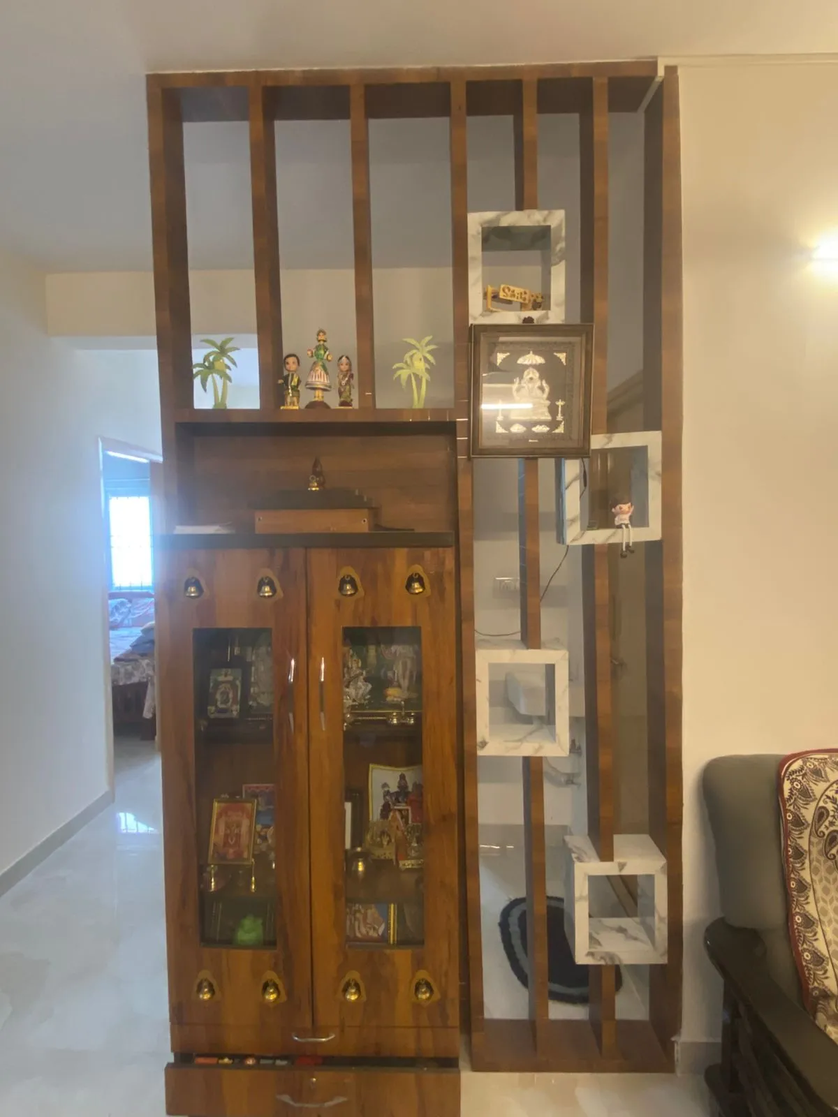 A wood pooja cabinet with brass bells and glass doors beside a slatted wood room-divider fitted with marble-inset display cubes.