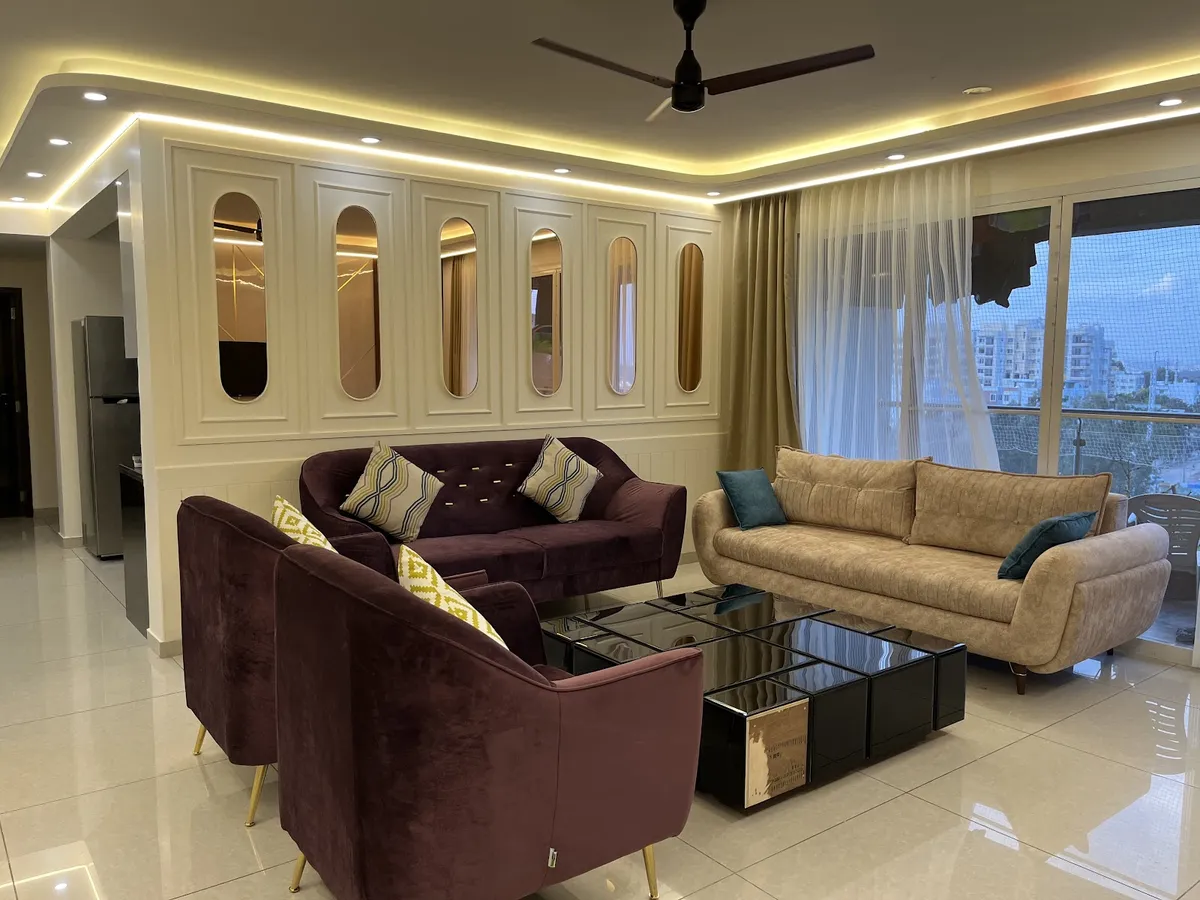 Living room where a cream moulded panel wall is punched with a row of capsule-shaped bronze mirror insets behind a plum velvet sofa and tub chairs