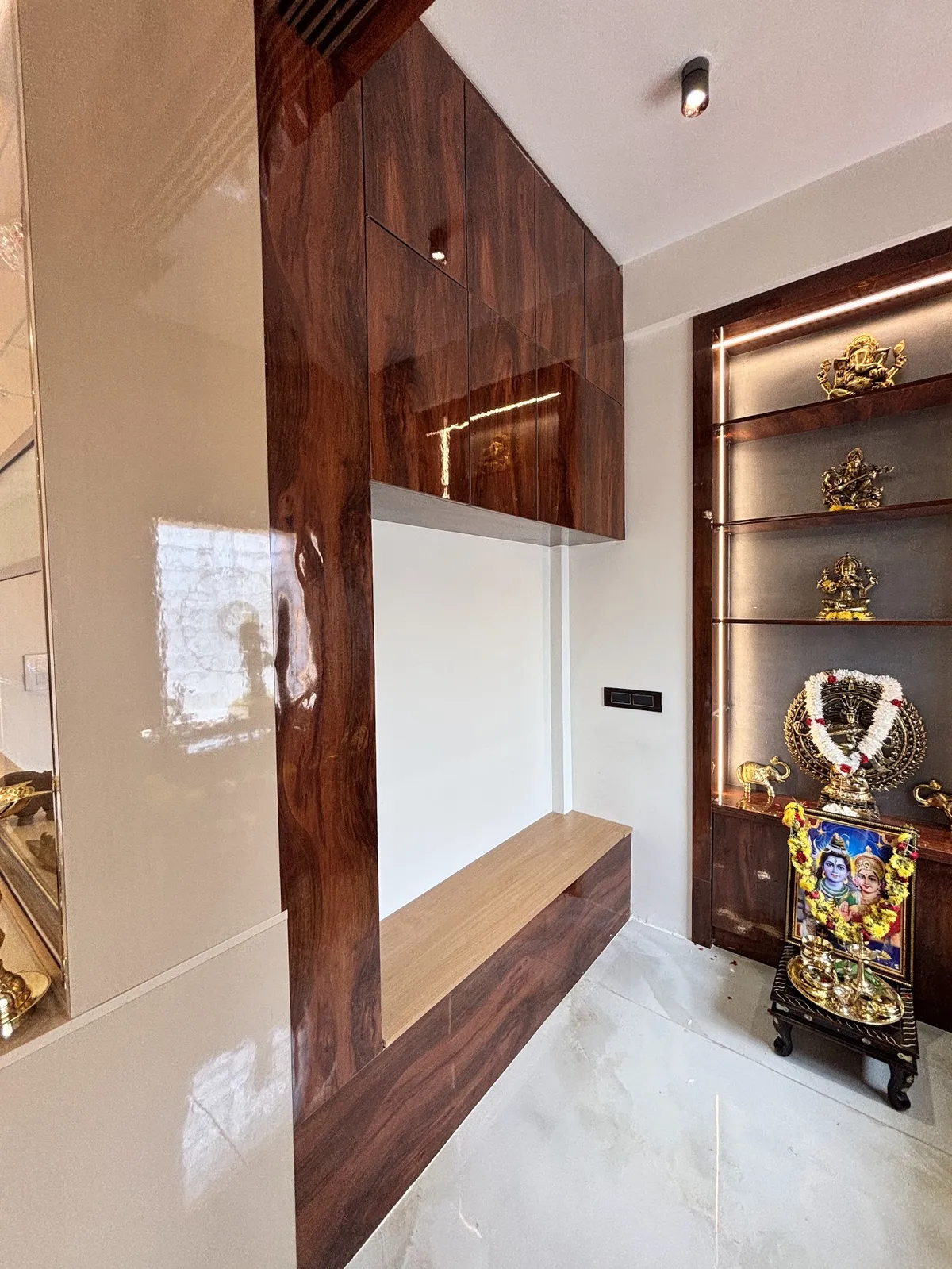 Backlit glass-shelf pooja unit in a dark wood-veneer frame displaying brass deity idols and a garlanded photo, beside a glossy wood-veneer wall and bench.