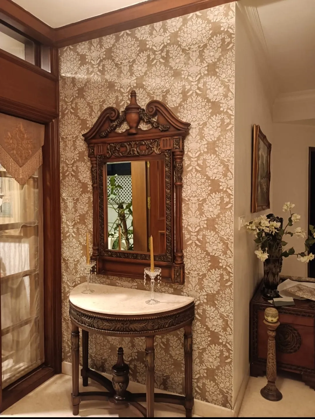 An entrance corner with a demilune marble-top console table below an ornately carved wood mirror, set against a gold-and-cream damask wallpaper