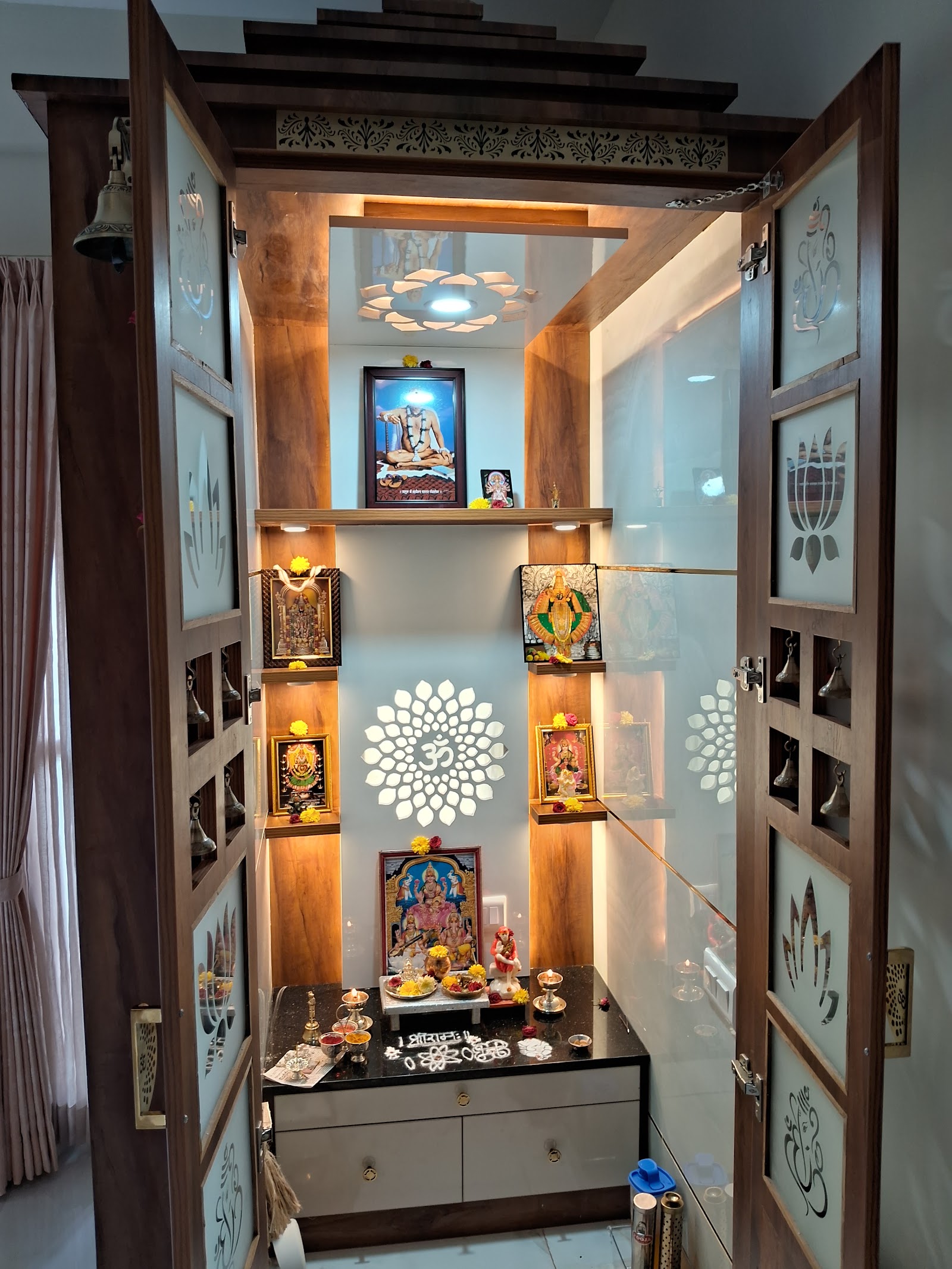 Dedicated pooja room with a carved wood temple unit, glass doors etched with deity motifs, a backlit white jaali Om medallion on the rear wall