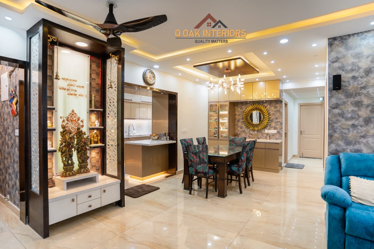 Real open-plan apartment showing an ornate white-and-wood pooja unit beside an open kitchen, a floral-upholstered dining set under a crystal chandelier
