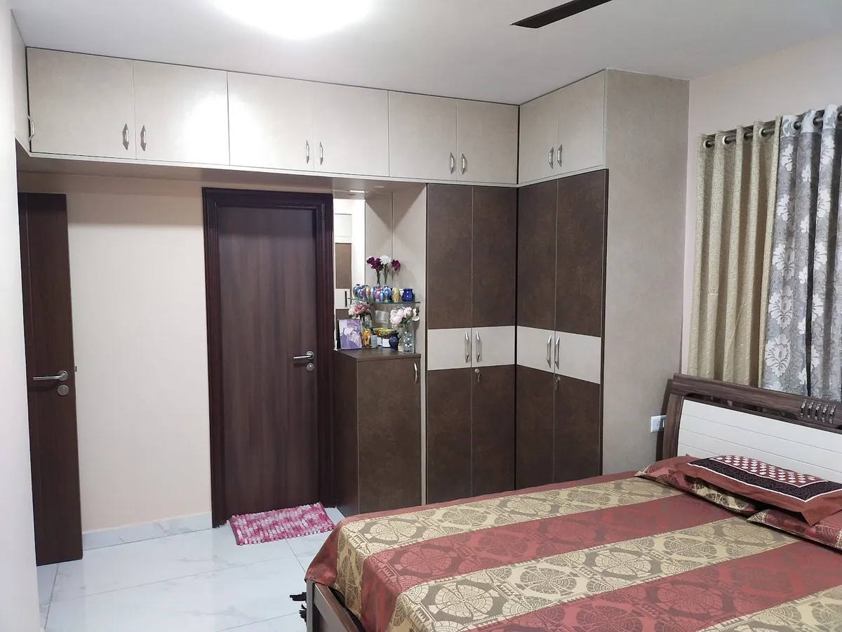 Lived-in bedroom where a two-tone brown-and-cream laminate wardrobe with loft shutters wraps into a corner dressing unit topped with glass shelves of coloured