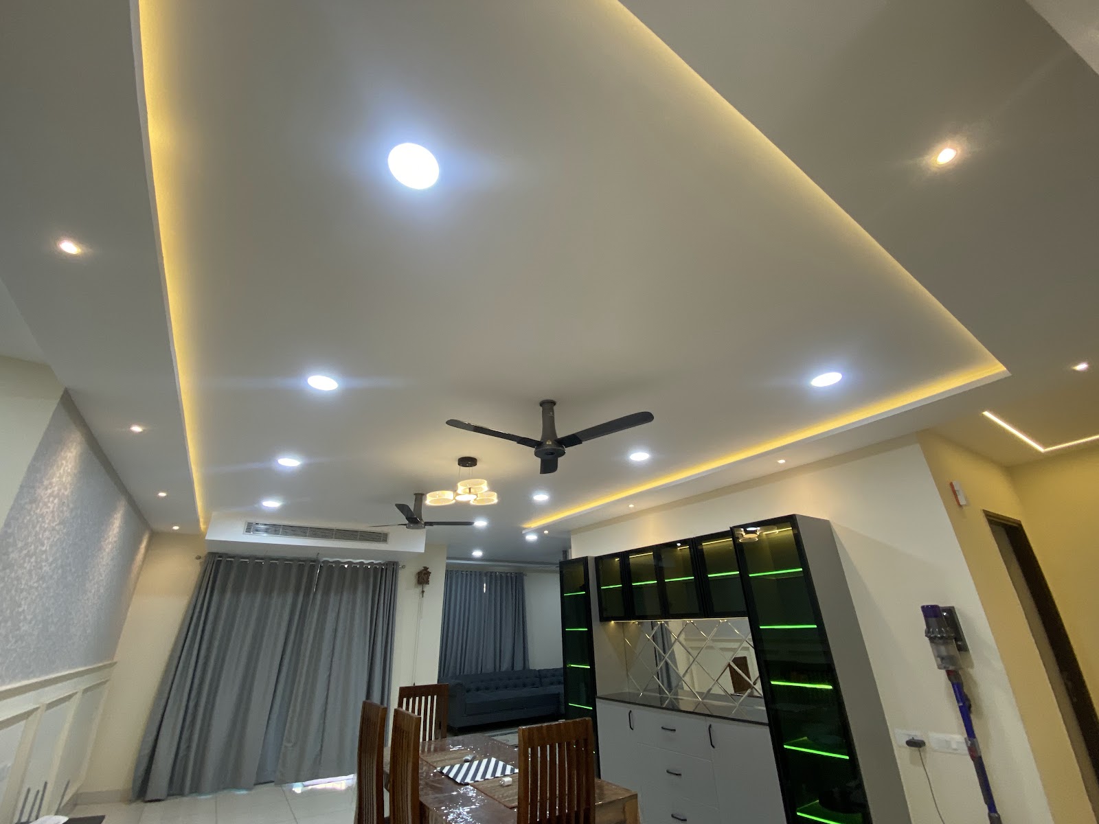 Dining-living space shot from a low upward angle dominated by a layered cove-lit POP ceiling with two fans, grey floor-length curtains behind