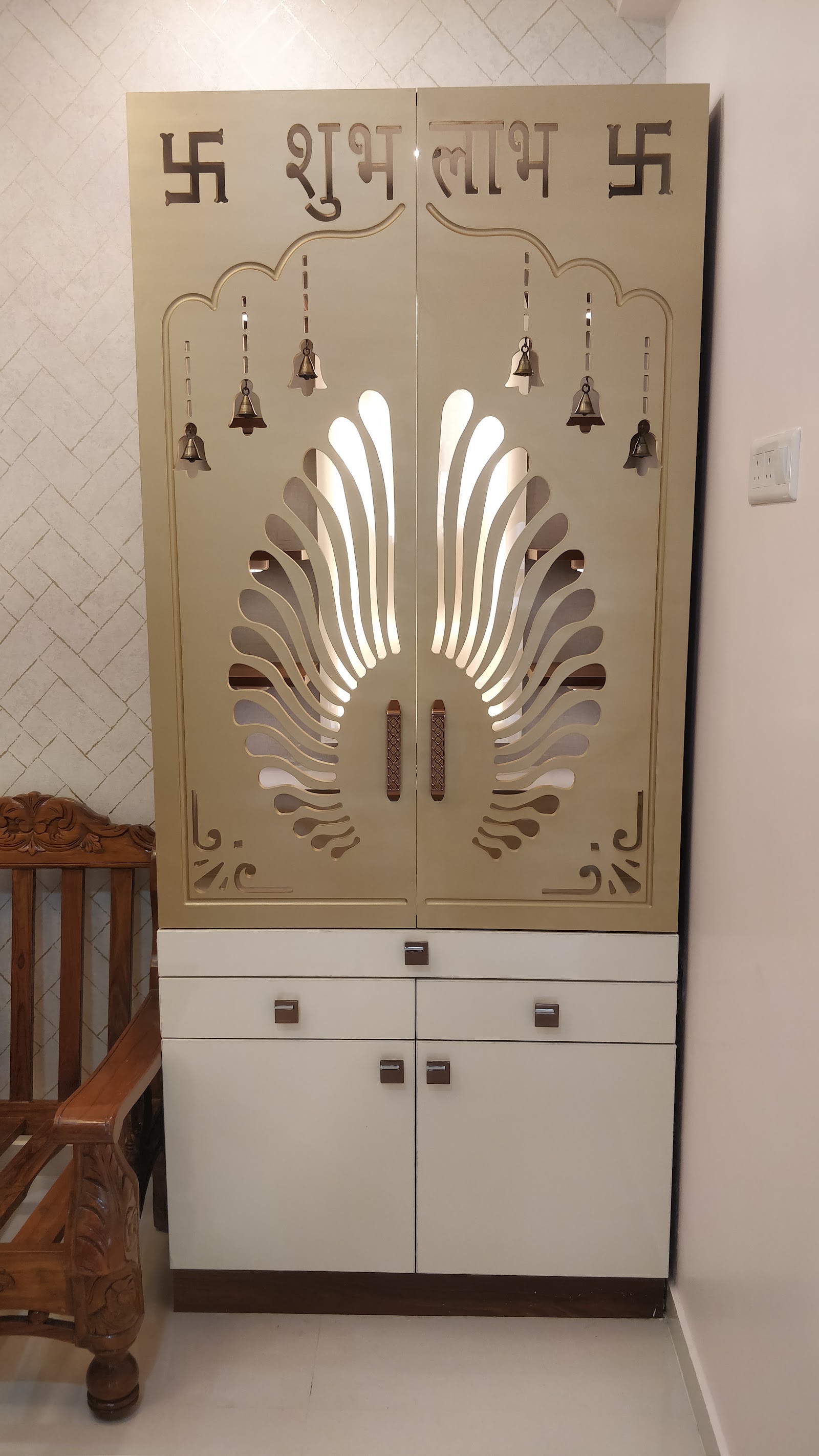 A cream-and-gold pooja unit with a laser-cut peacock-feather jaali door lit from behind, flanked by hanging bells and a Shubh Labh swastik motif.