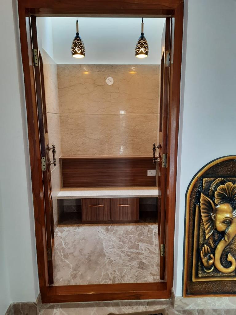 Pooja room seen through open wood-framed doors, with marble-clad walls, a built-in altar shelf with drawers, two pendant lights