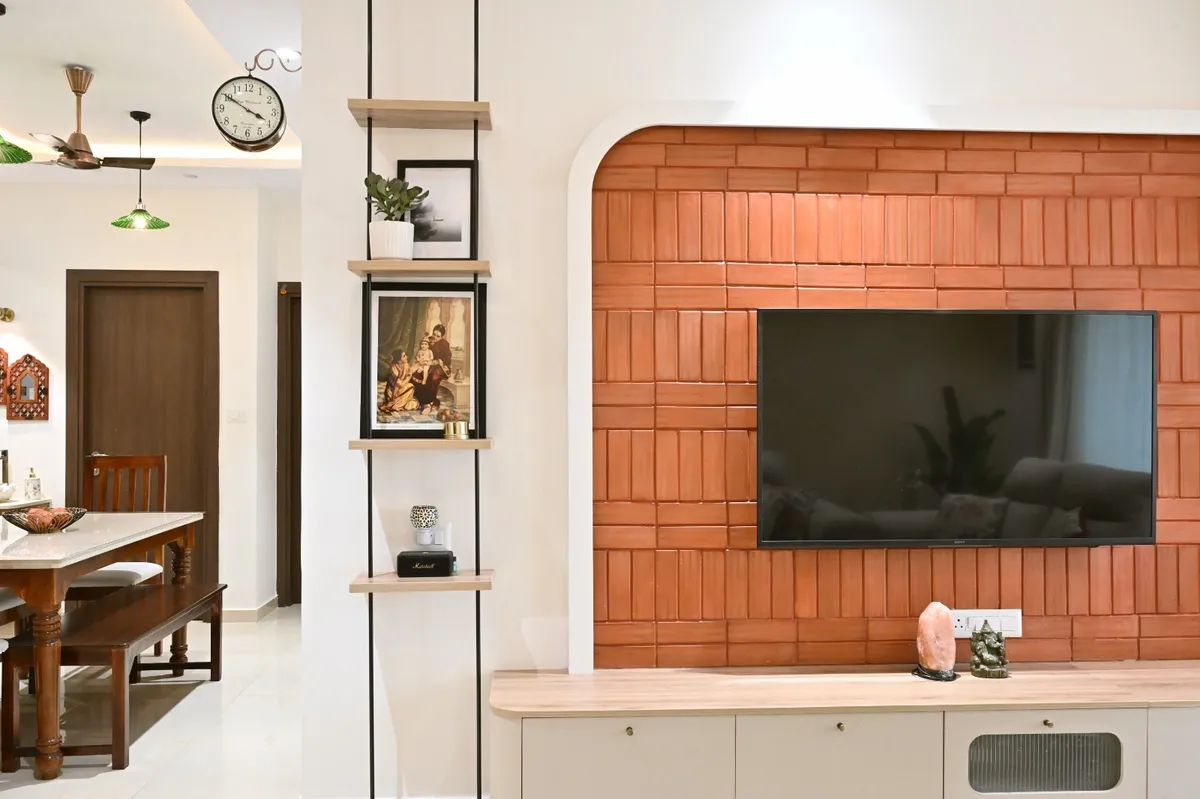 Terracotta brick-tile TV wall set inside a white arched niche, with a light-oak floating console and a suspended four-tier shelf carrying a Ravi Varma print
