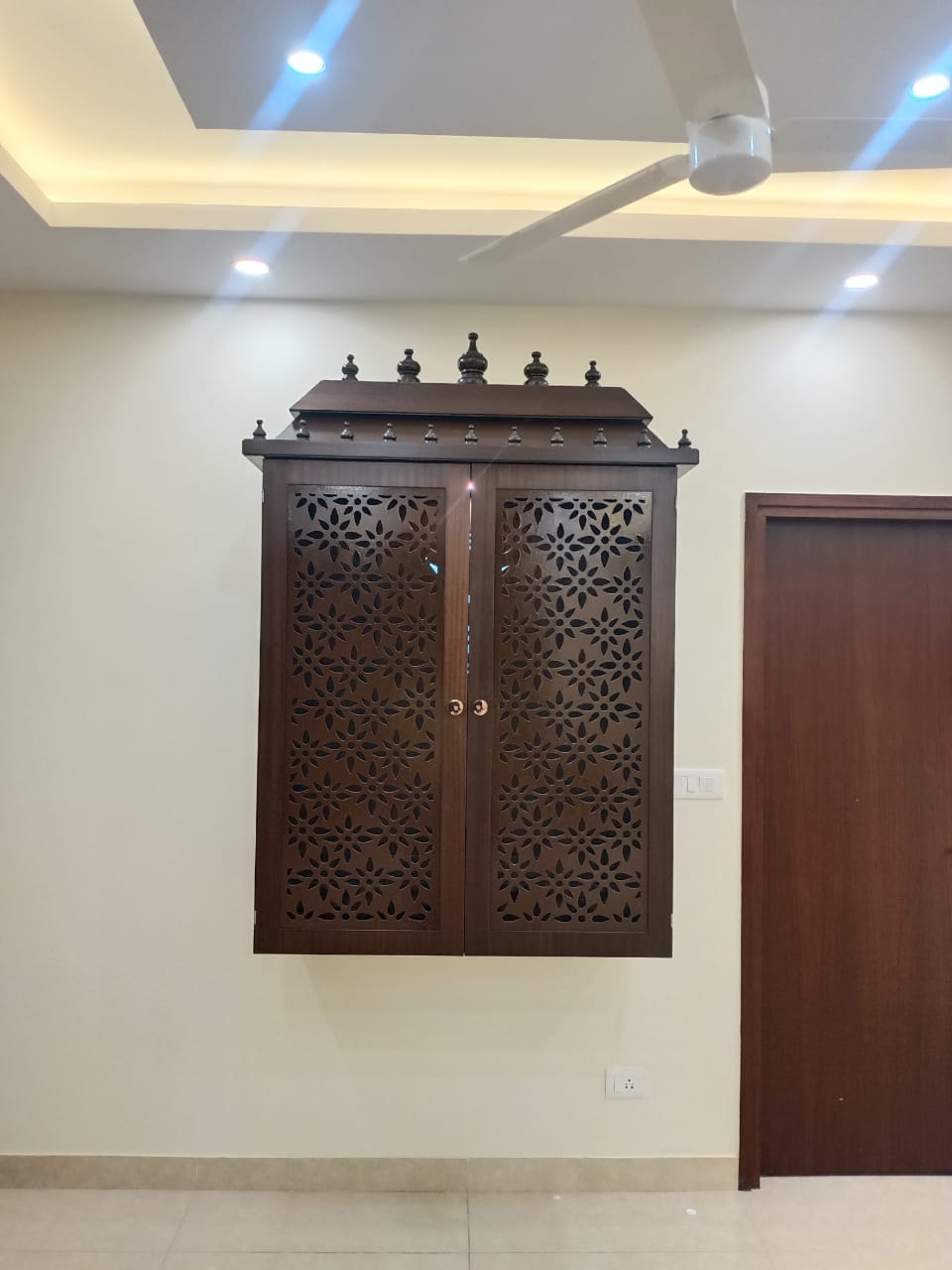 A wall-mounted wooden mandir unit with intricate floral jaali-cut doors and temple-style finials, set against a plain wall below a cove-lit ceiling