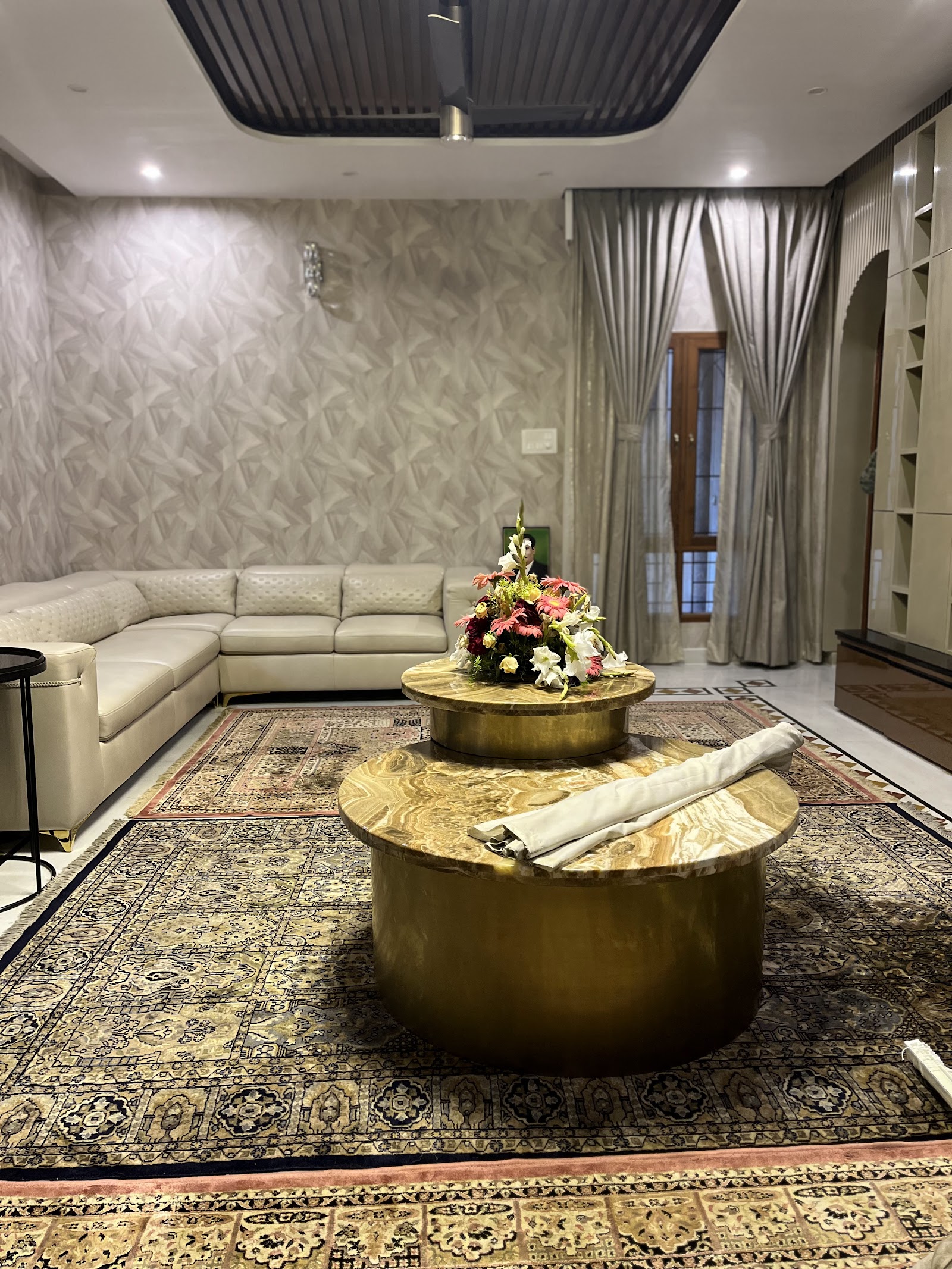 A living room with a cream leather sectional, layered Persian-style rugs, twin round onyx-and-brass coffee tables, silvery silk drapes
