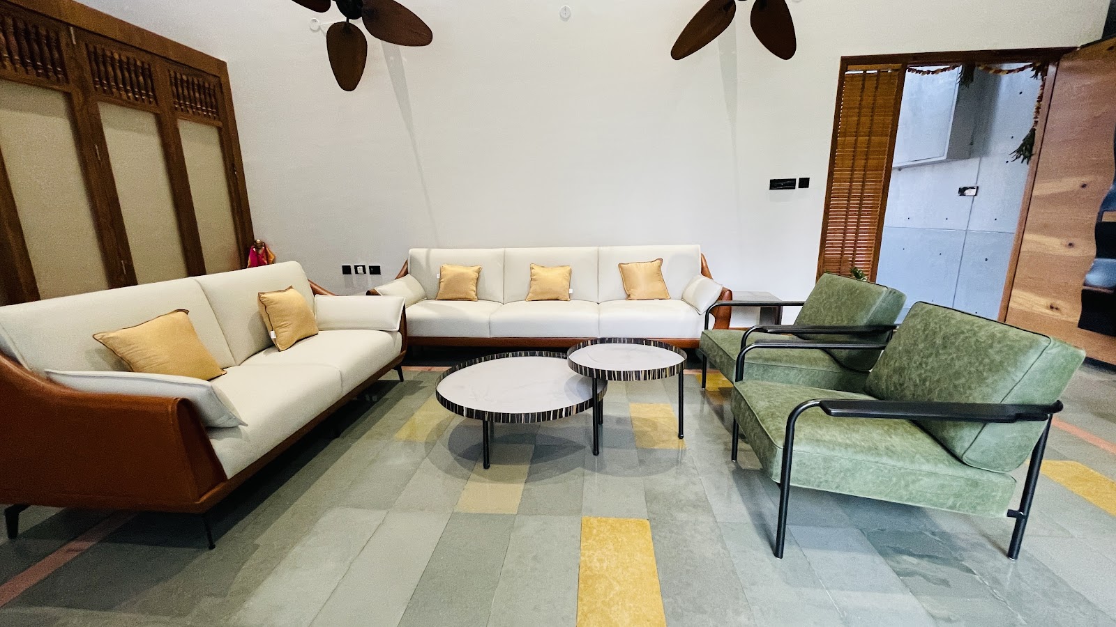 Living room with tan leather and cream sofas, olive-green mid-century armchairs, and twin round marble coffee tables on a stone floor with ochre-yellow inlay