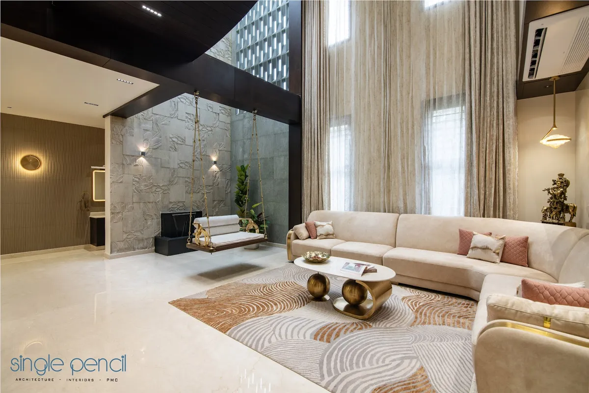 Double-height living room where a curved cream velvet sectional faces a brass-chain jhula hung against a carved leaf-relief stone wall