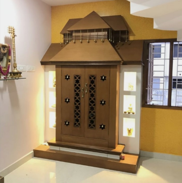 Pooja room with a temple-shaped wooden mandir unit featuring star-cutout jaali doors and lit display niches, set against a mustard-yellow textured wall.