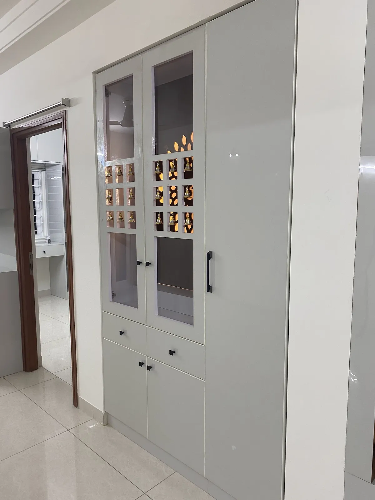 Newly installed floor-to-ceiling pooja cabinet in pale sage-grey shutters, its glazed upper doors framing a backlit square-grid jaali hung with nine brass