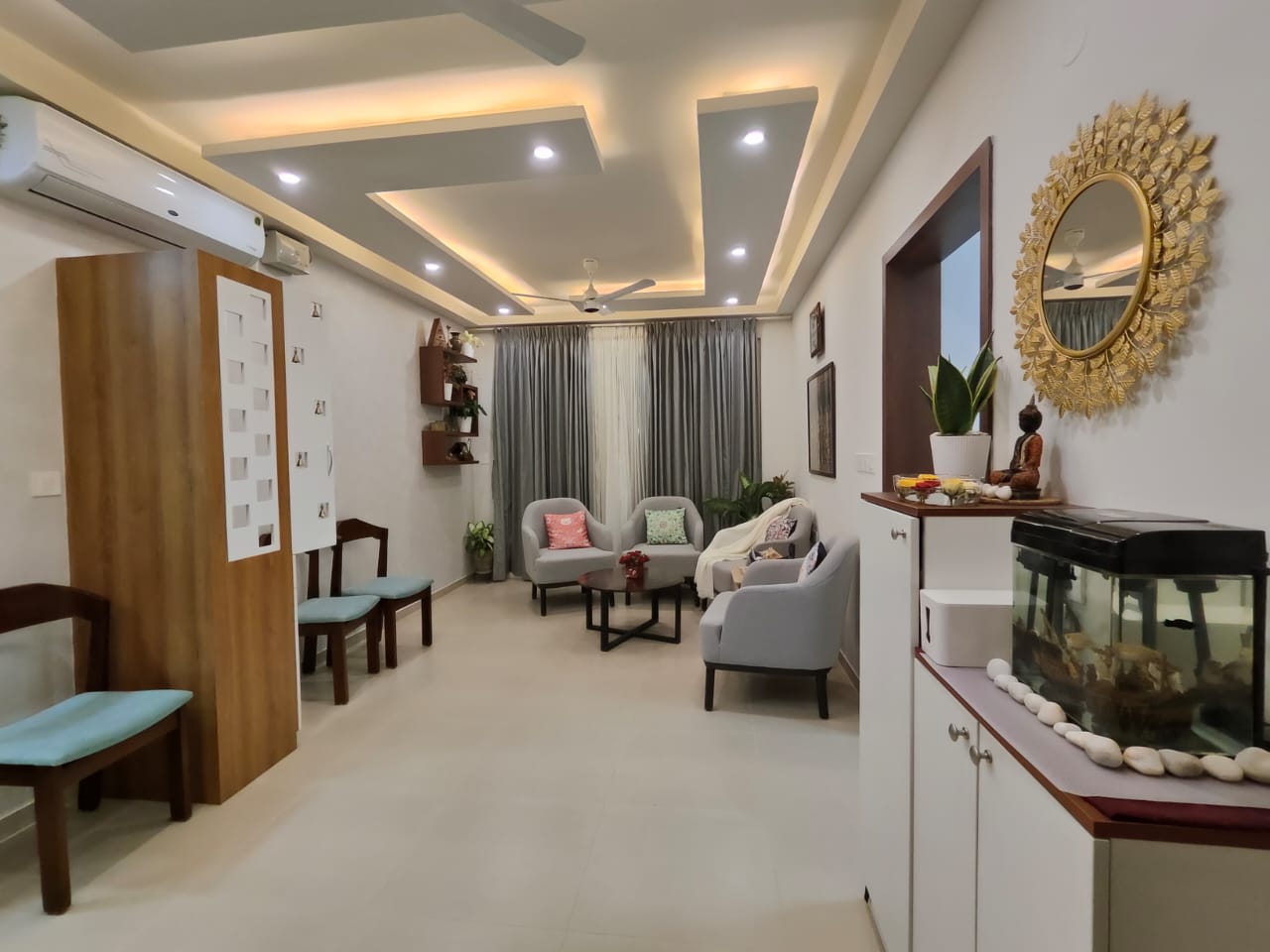 Living room with a multi-tier cove-lit false ceiling, grey armchairs around a black coffee table, and a gold sunburst mirror above a styled console.