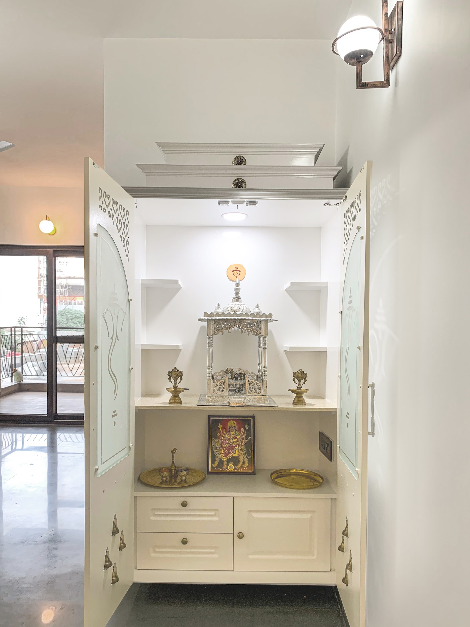 Built-in pooja unit with arched frosted-glass cabinet doors, hanging brass bells, a carved silver temple, and brass ritual items on a marble-effect shelf.