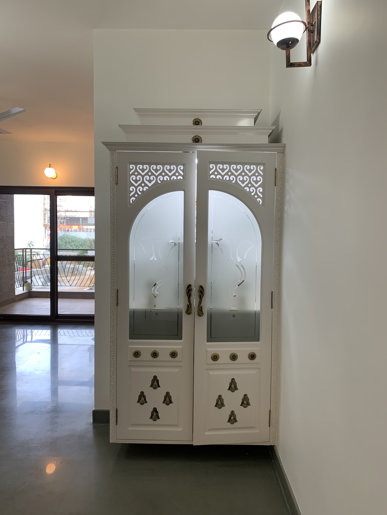 Free-standing white pooja unit with arched frosted-glass double doors, a carved jaali crown and brass bell-motif hardware