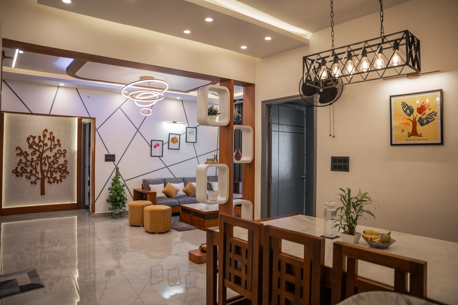 Open-plan entrance, living and dining area: a wooden tree wall-art foyer, grey sofa with mustard ottomans under a ring chandelier