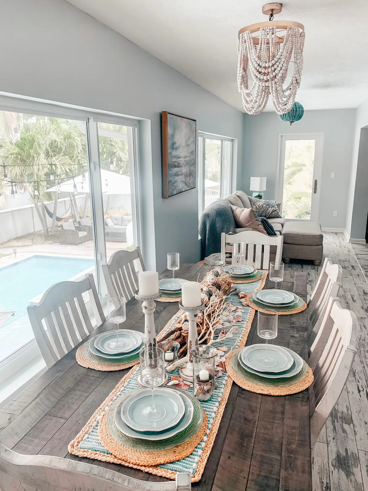 Weathered plank dining table laid with sea-green glass chargers on jute mats and a driftwood-and-shell runner