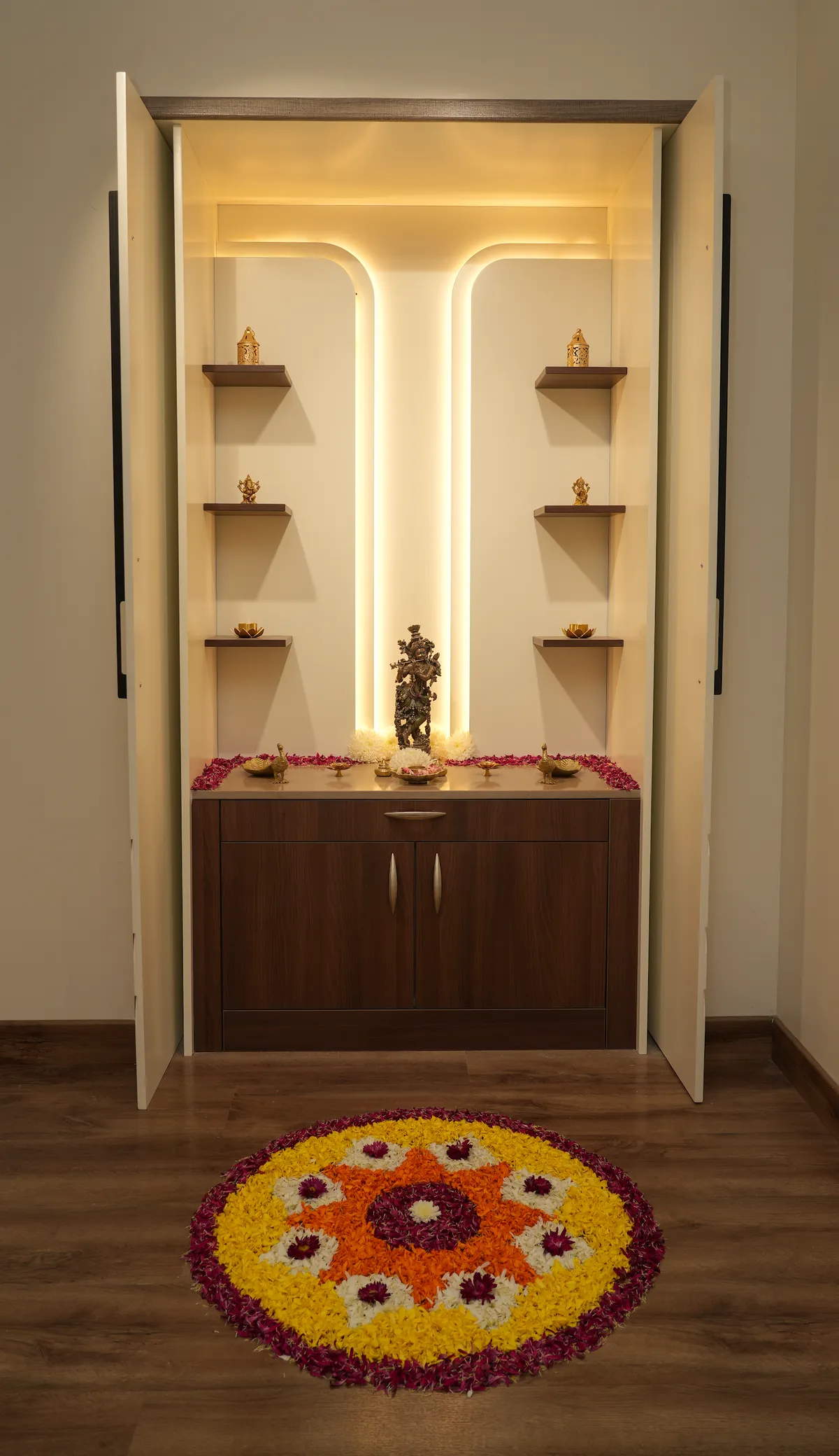 Pooja unit set in a niche behind full-height ivory shutters, a curved warm LED profile framing a brass Krishna idol on a wenge counter edged with rose petals
