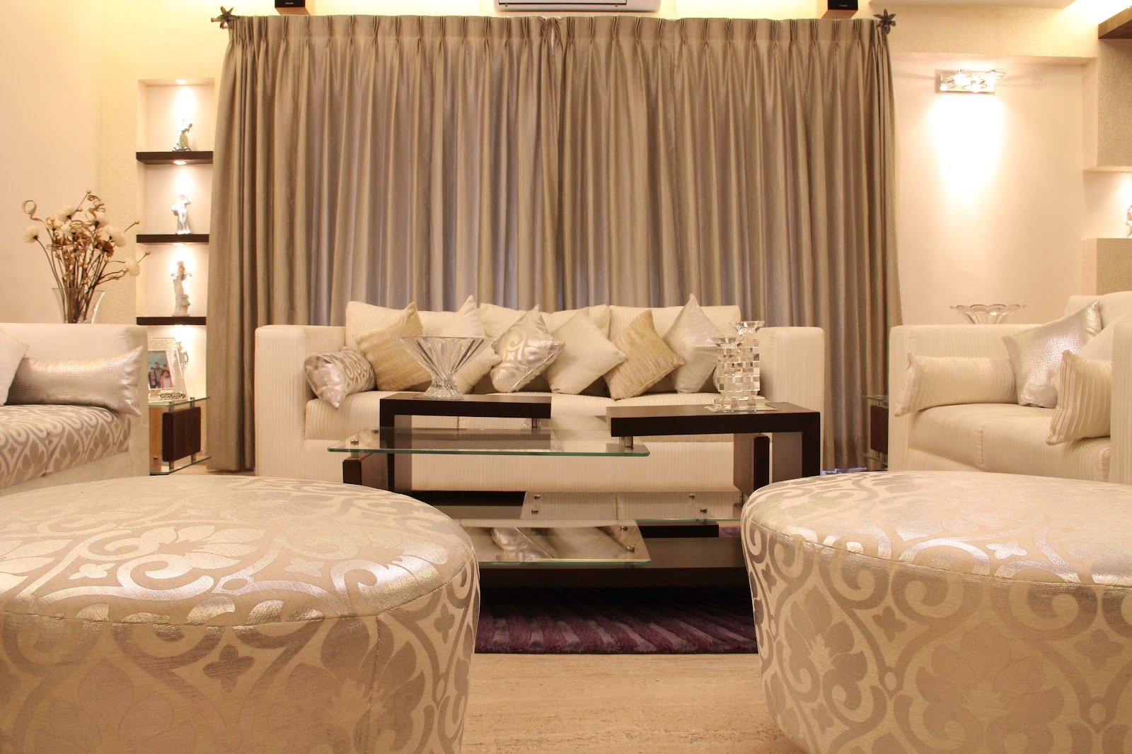 Cream and champagne living room with damask-patterned round ottomans, a glass-and-wood tiered coffee table