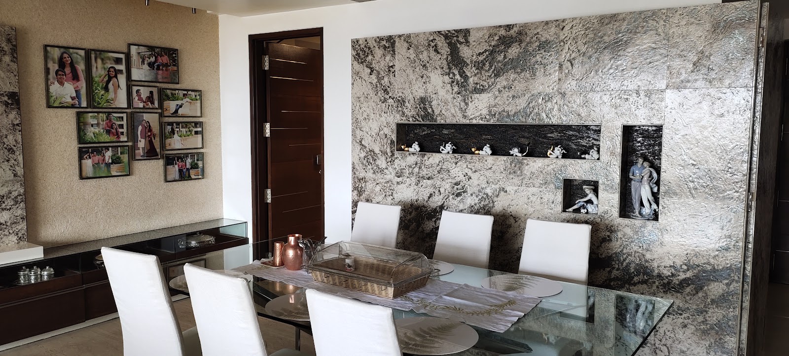 Dining area with a glass table and white leatherette chairs against a textured stone feature wall with lit display niches and a family photo gallery