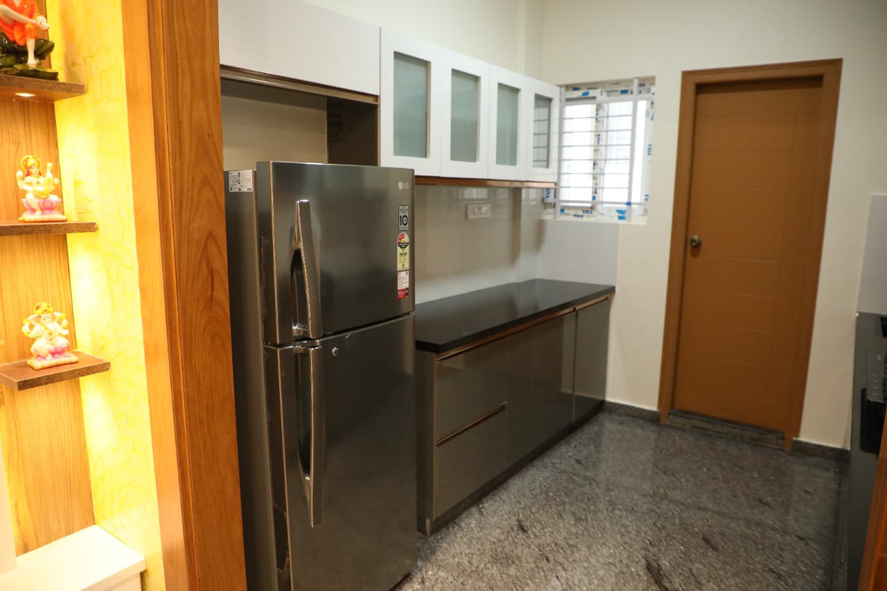 Galley kitchen with a stainless steel refrigerator, dark gloss cabinetry and a black granite counter, with a small lit pooja shelf just outside the doorway