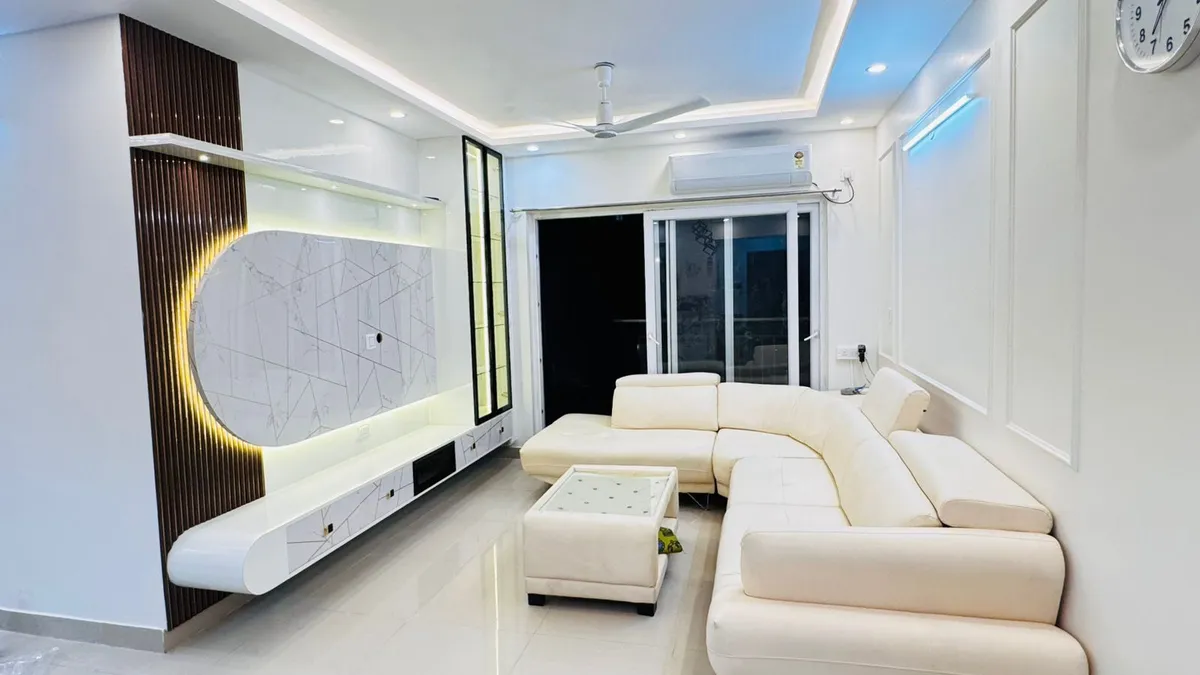 L-shaped cream sectional sofa facing a curved fluted-wood TV unit with a backlit oval marble-pattern panel, in a living room with cove ceiling lighting