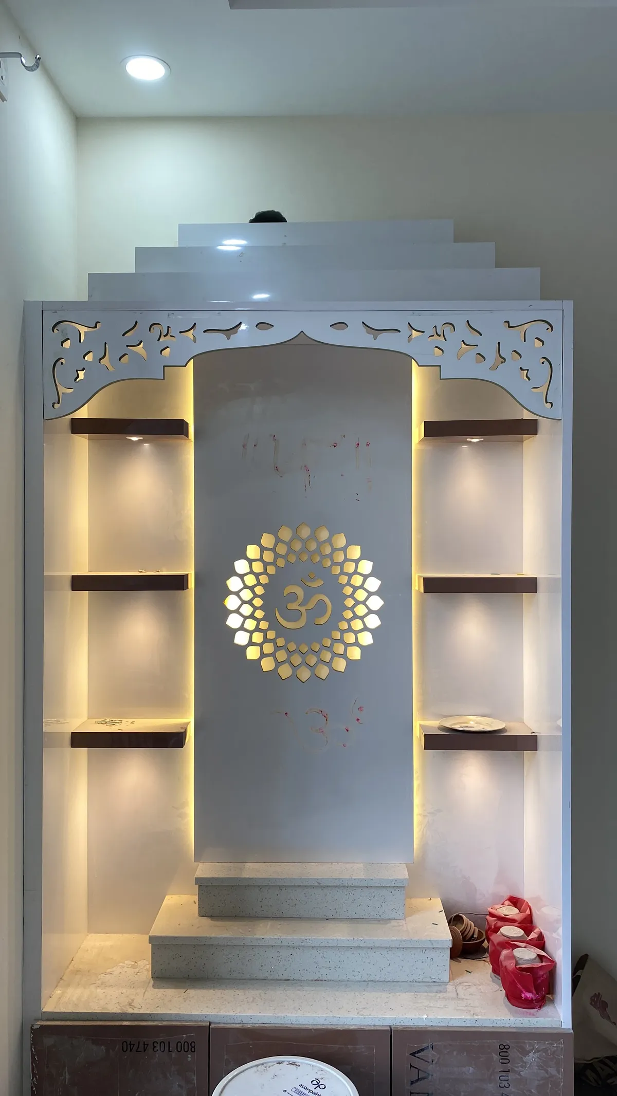 Pooja unit with a white carved jharokha-style arch, a backlit gold Om medallion on the mandir backwall, illuminated display shelves on either side
