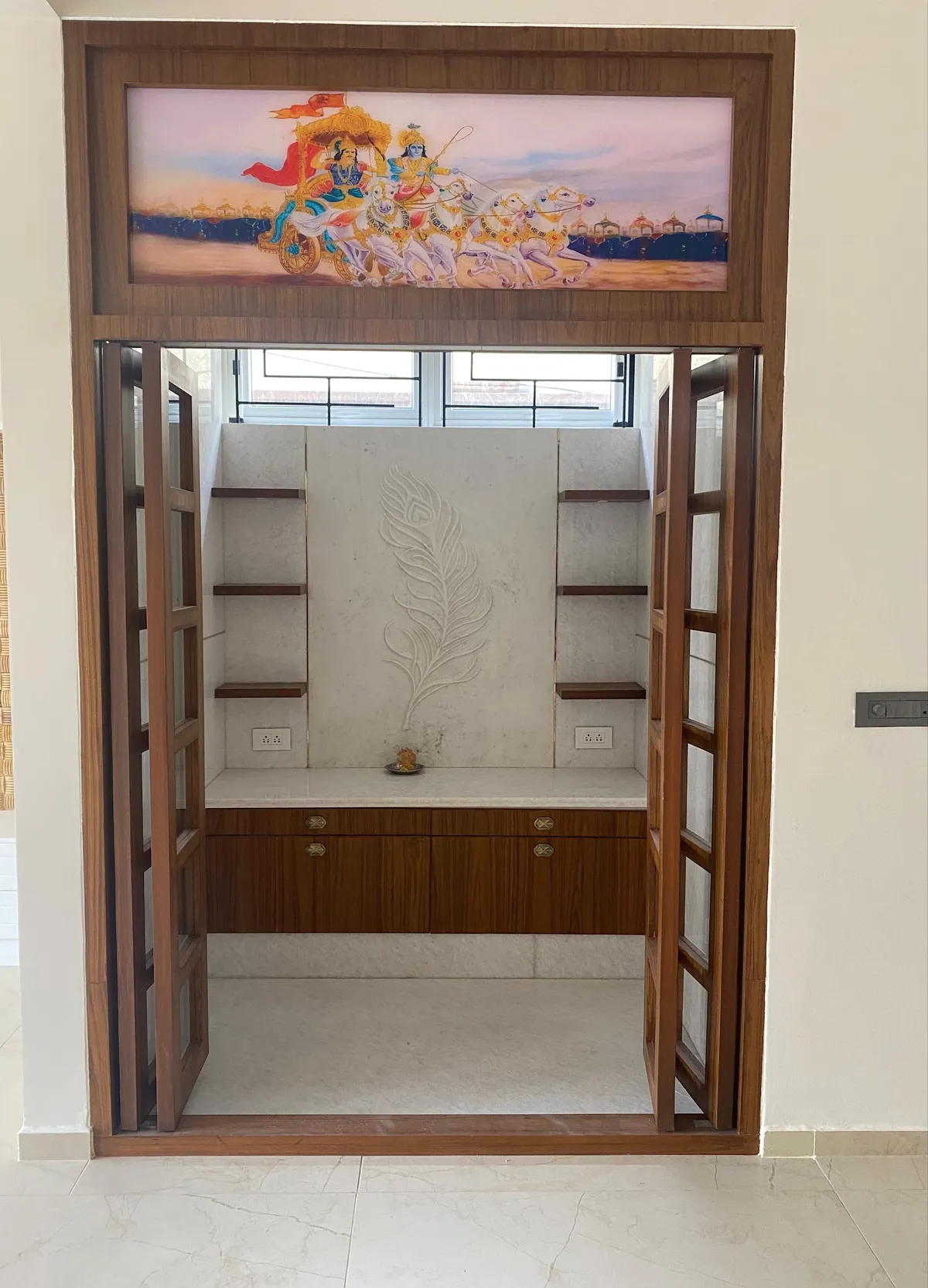 A pooja alcove behind folding glass-and-wood doors, with a hand-carved marble peacock-feather backdrop and a backlit mythological mural above the lintel.