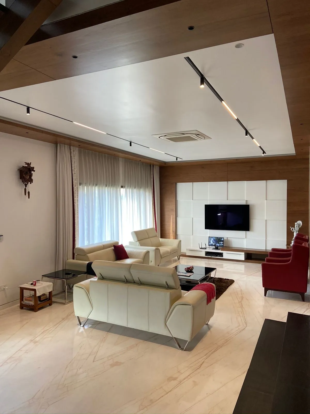 Large living room on cream veined marble floors with cream leather recliner sofas facing a TV wall of white upholstered square pads set into wood veneer