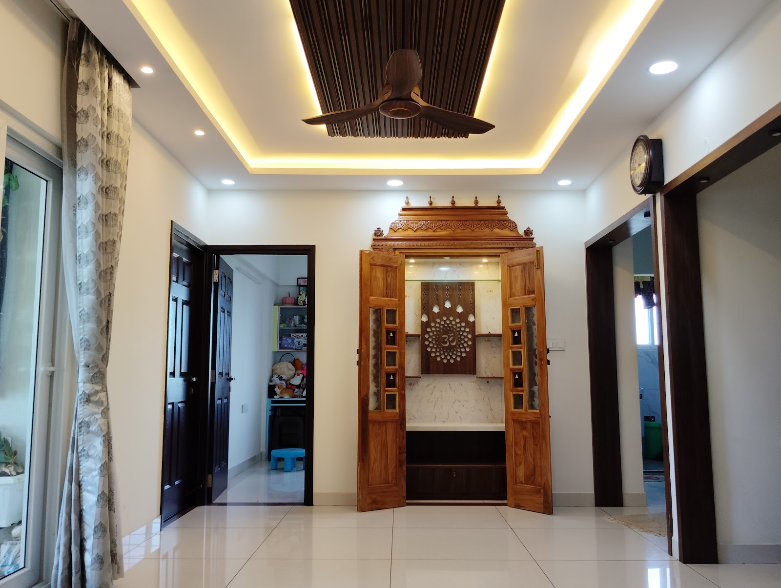 Wall-recessed pooja unit with an intricately carved teak door frame, a brass Om motif jaali panel, and a marble backdrop under a cove-lit ceiling.