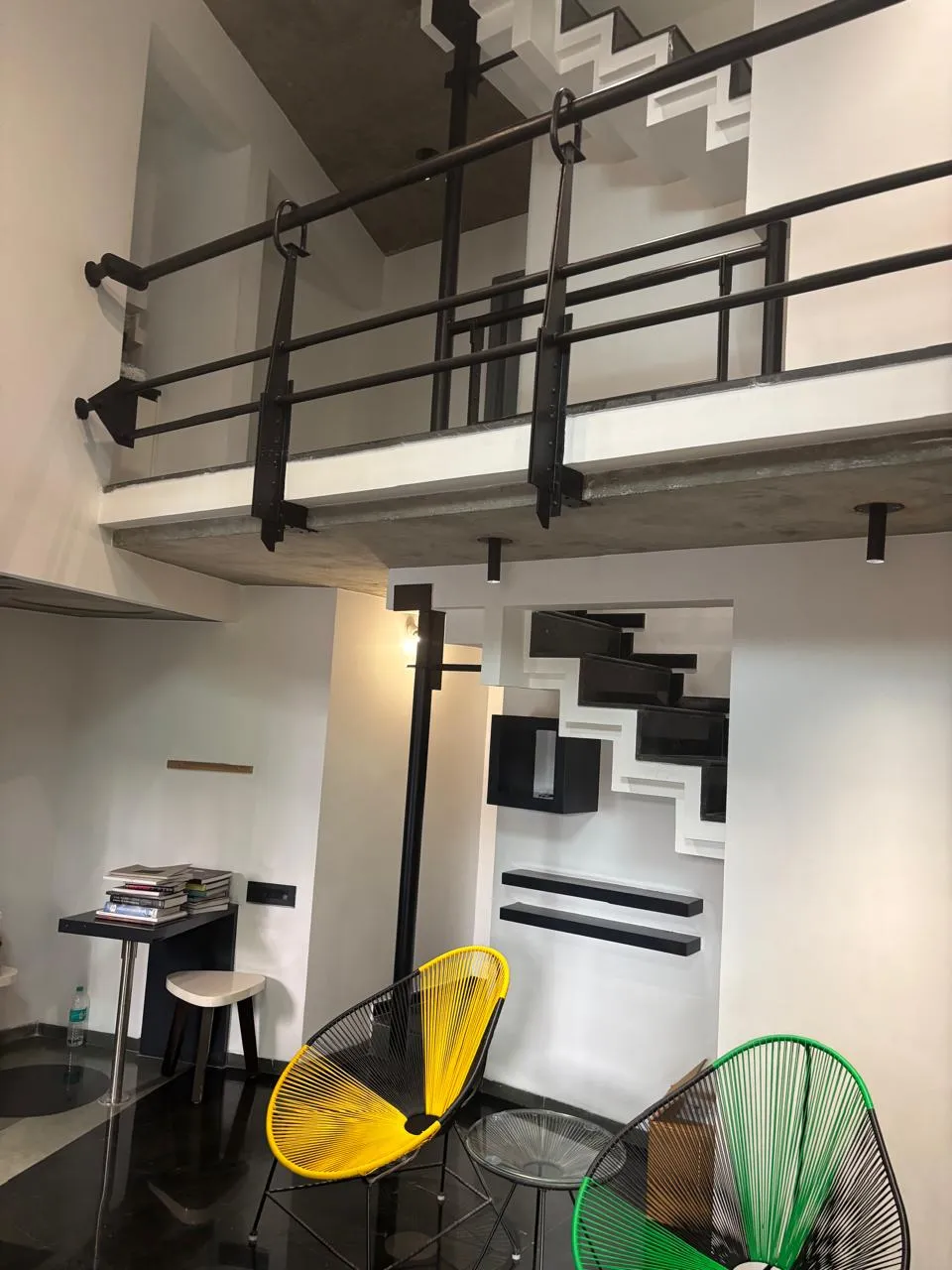 Double-height industrial loft with an exposed concrete mezzanine, black pipe railings, and yellow and green wire-frame accent chairs below the stair.