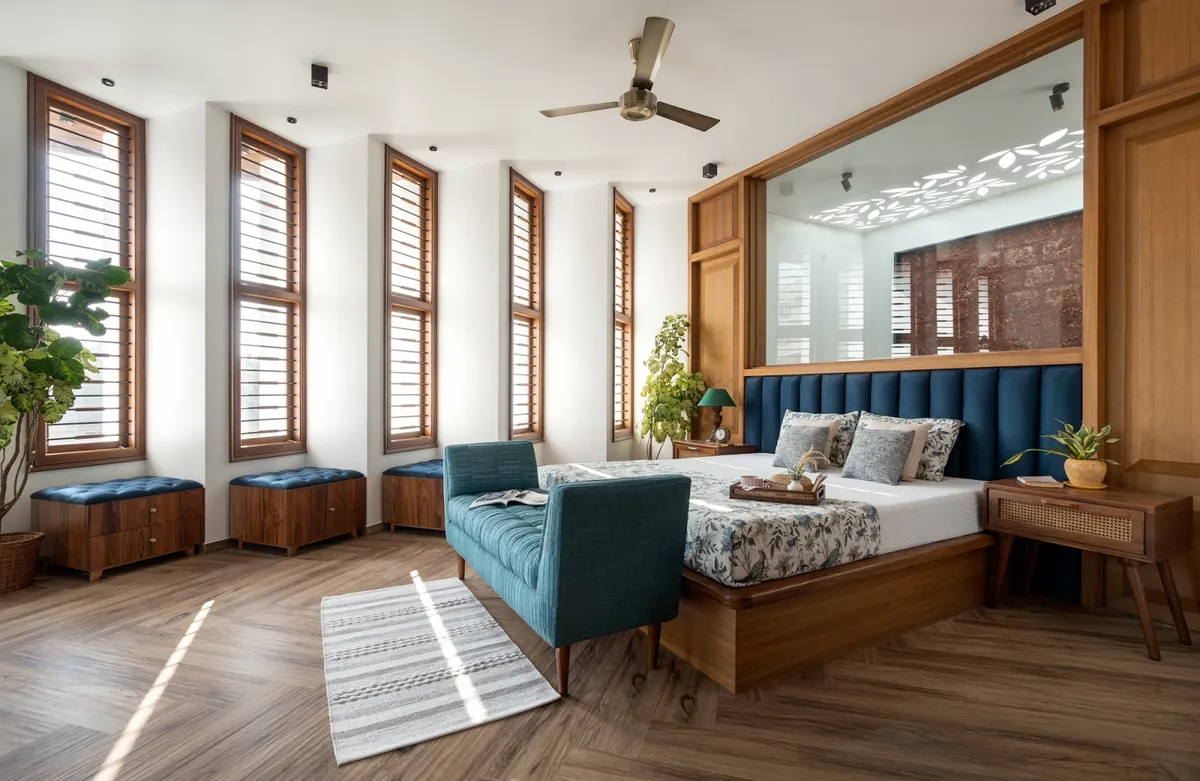 A wood-floored bedroom with a tall louvred window wall, a backlit jaali screen behind a teal channel-tufted headboard