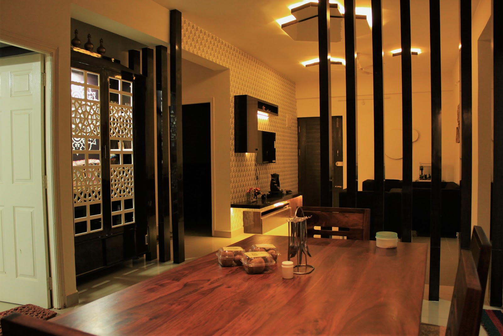 Real photo of a dining room with a solid-wood dining table in the foreground, a black jaali-fronted pooja cabinet to the left