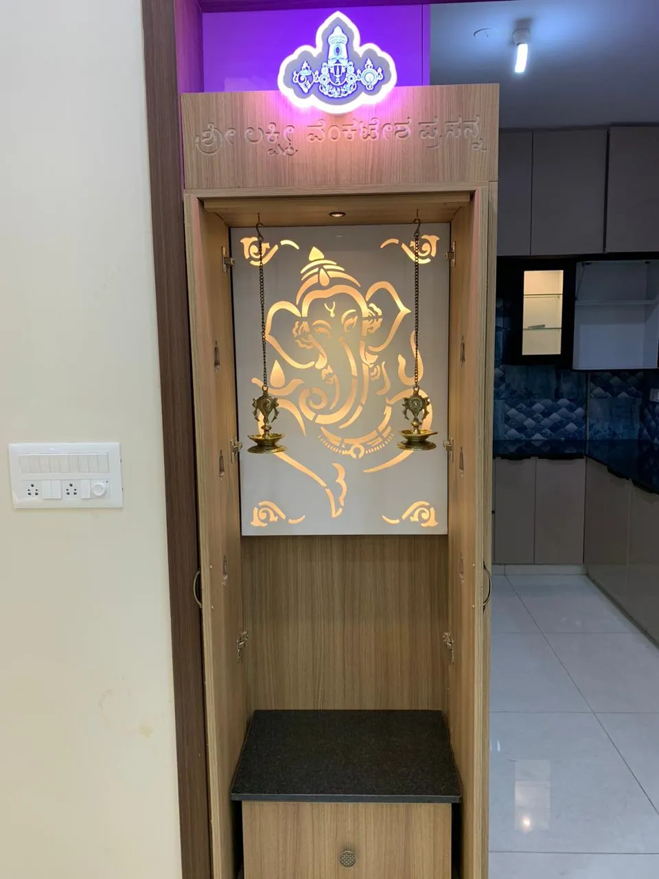 Compact pooja unit with a backlit laser-cut Ganesha jaali panel, wood cabinetry and a granite-top base shelf.