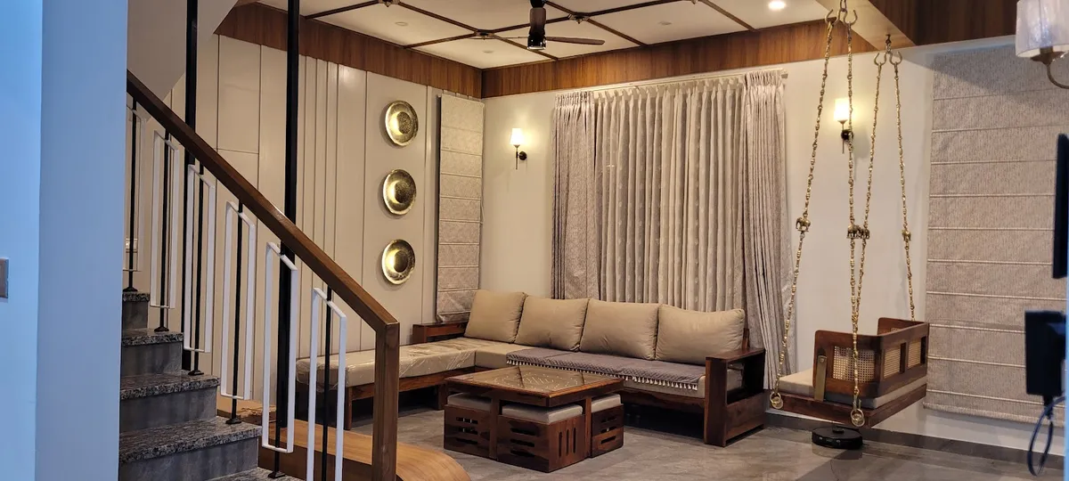 Living room with an L-shaped wood-frame sofa, a brass-chained hanging swing, three brass wall plates, and a wood-panelled coffered ceiling.
