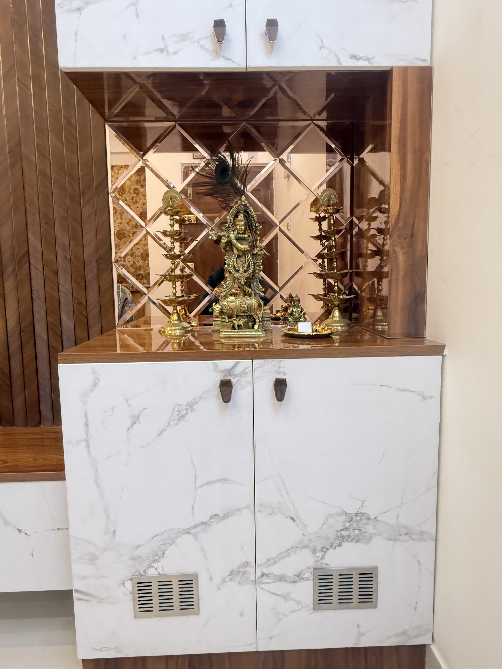 Wall-mounted pooja unit with a diamond-pattern mirror backsplash, marble-effect laminate cabinet doors, and brass idols and diyas on the wood-laminate shelf.
